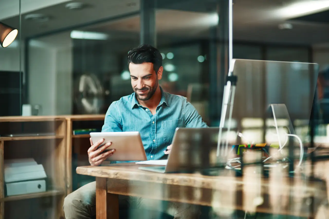 A imagem apresenta um homem jovem vestindo roupa social despojada, e que está com um tablet em uma das mãos. Ele está sentado em frente a uma mesa com seu notebook. O tema do artigo é empreendedorismo tecnológico.